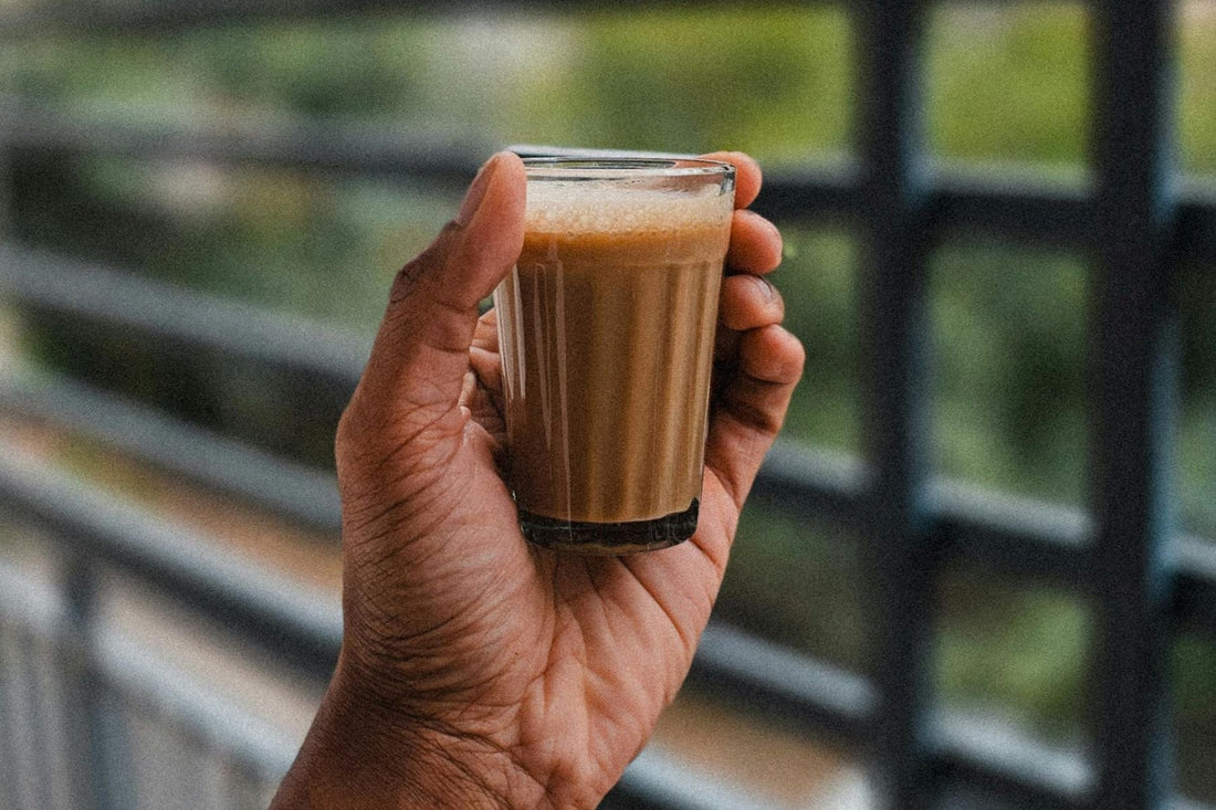 Close-up of a hand holding a small glass of creamy chai or milk tea, with a blurred outdoor railing and greenery in the background.