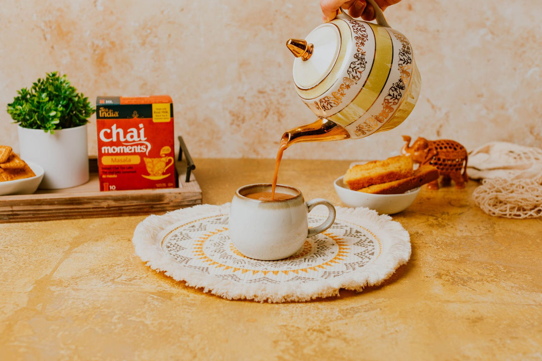 A teapot pouring hot chai into a cup, with Tea India Chai Moments and tea-time snacks arranged in a warm, cozy setting.