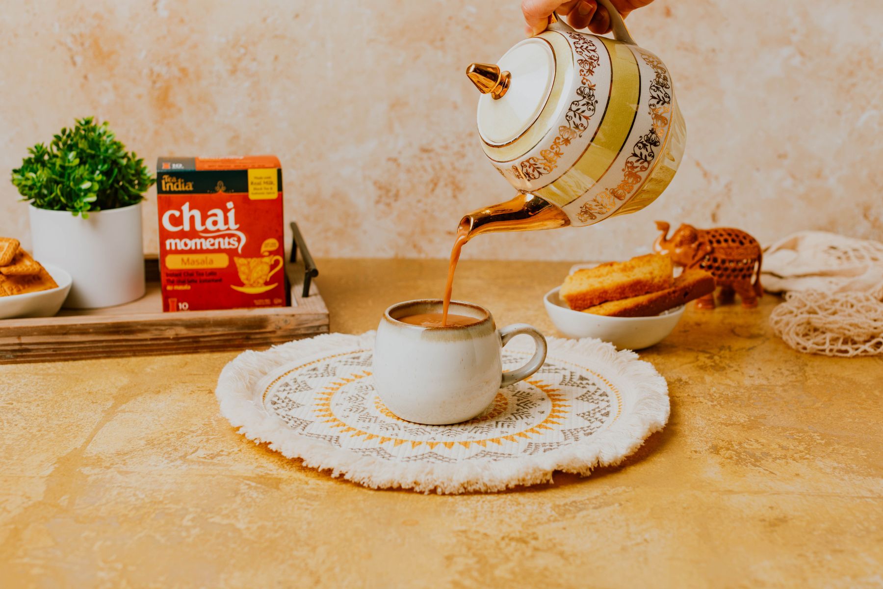 A teapot pouring hot chai into a cup, with Tea India Chai Moments and tea-time snacks arranged in a warm, cozy setting.