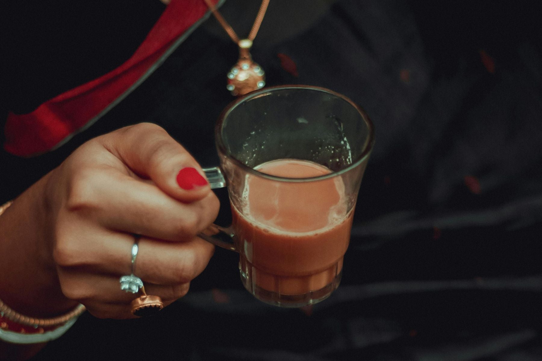 Hand holding a small glass cup of warm chai with red-painted nails and jewelry in a cozy low-light setting