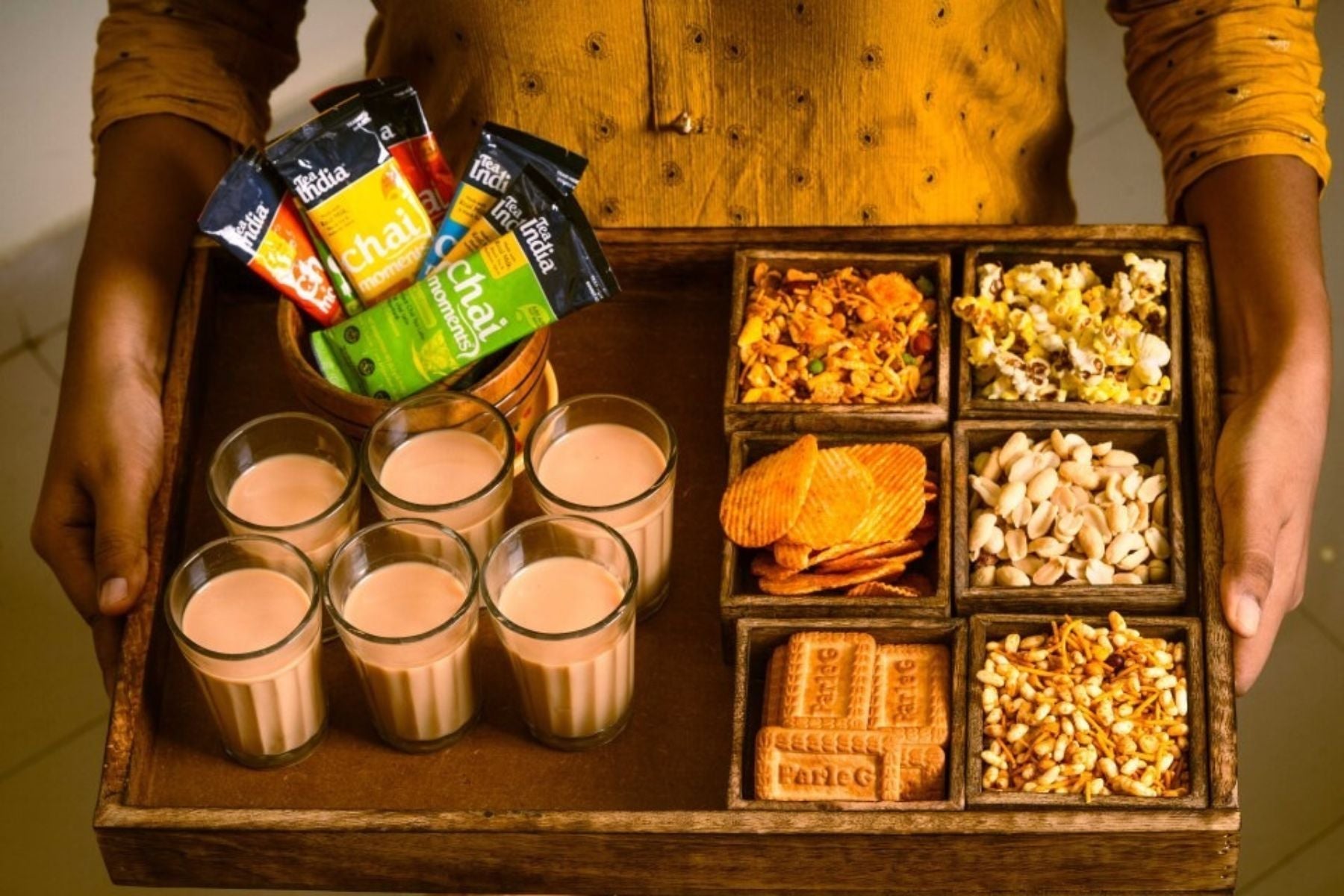 A hand holding a tray of assorted snacks, 6 glasses of tea and several chai moment sachets