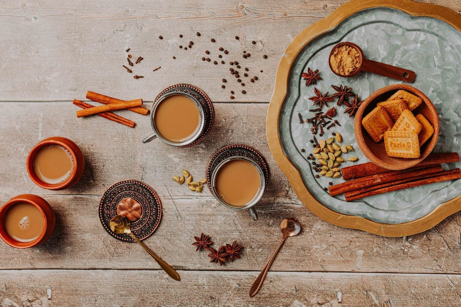 Four cups of tea alongside spices and biscuits on a plate.