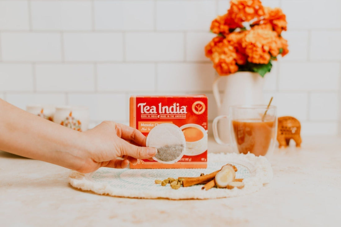 Hand holding a Tea India Masala Chai tea bag in front of the box with spices and brewed chai