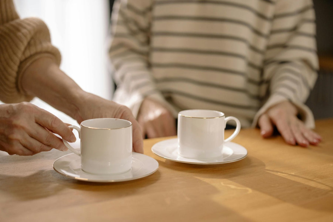 Two white cups of tea being placed on a table as someone sits across, creating a warm shared moment