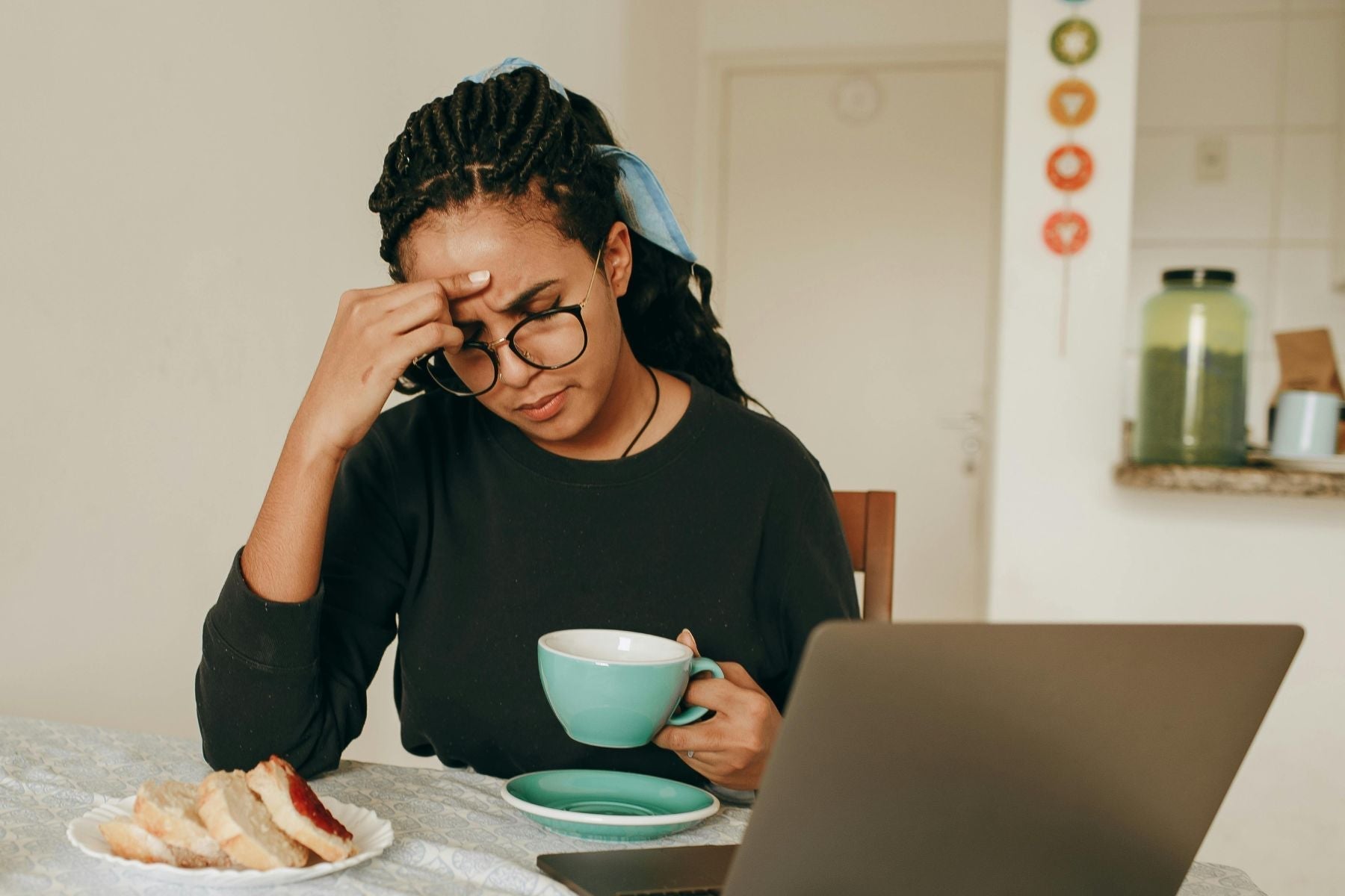 Woman looking stressed while holding a cup of tea, working on a laptop at the table