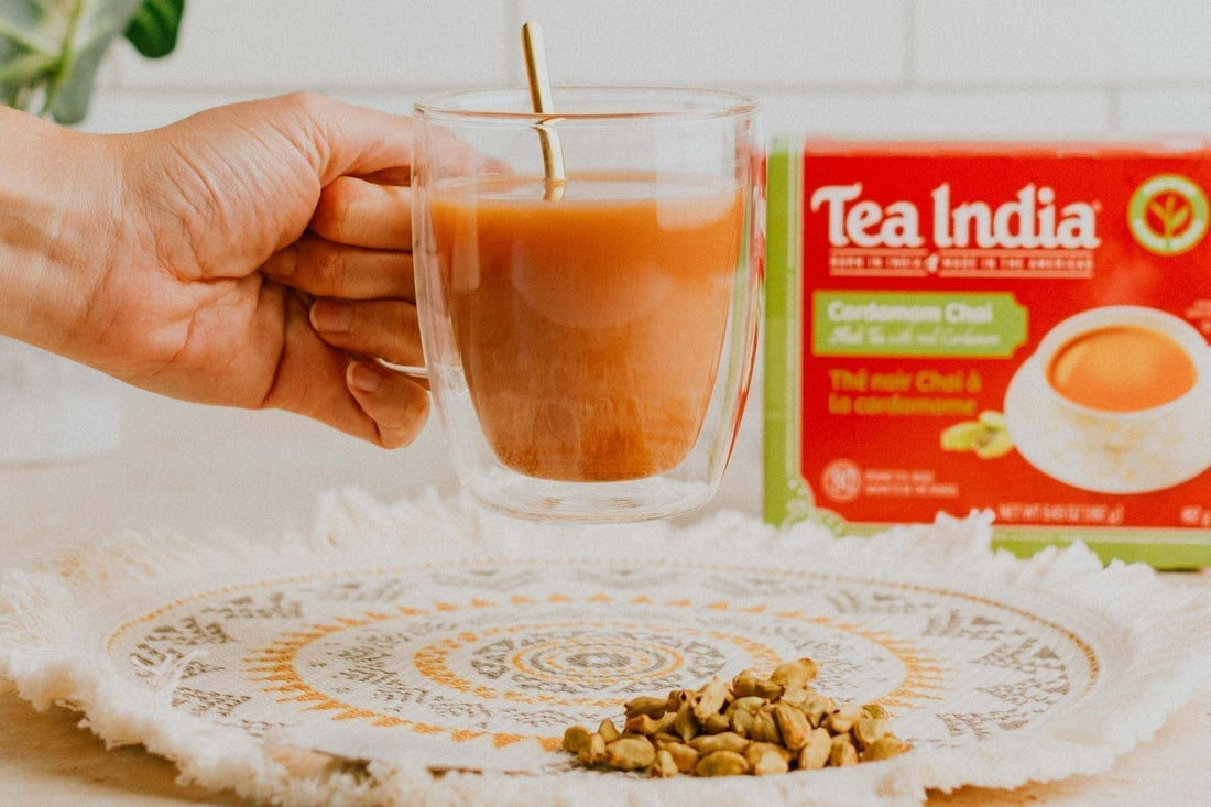 Hand holding a glass cup of chai with Tea India Cardamom Chai box and crushed cardamom in the background