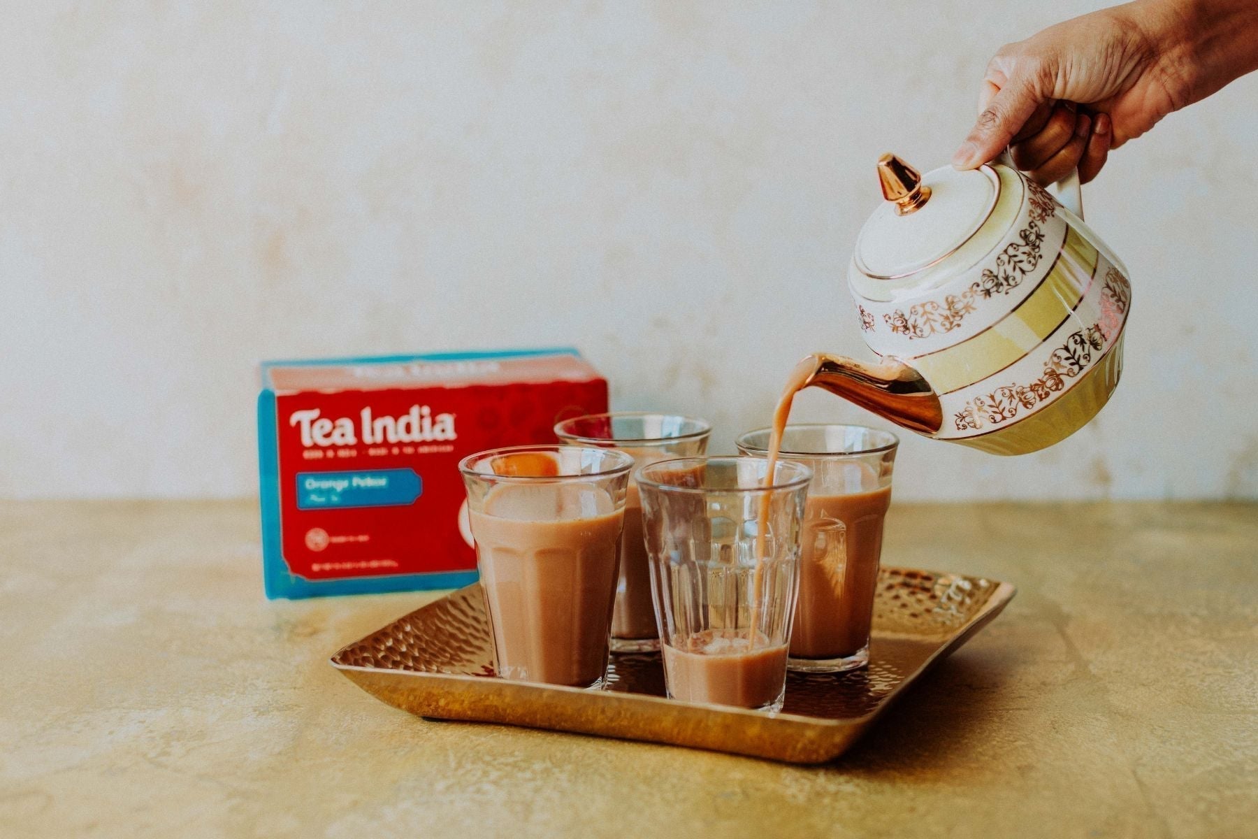 Tea being poured from a kettle into multiple glasses with Tea India box in the background