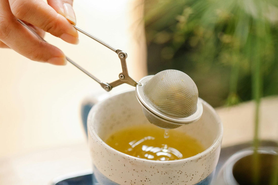 Hand lifting a tea infuser above a cup of freshly brewed green tea