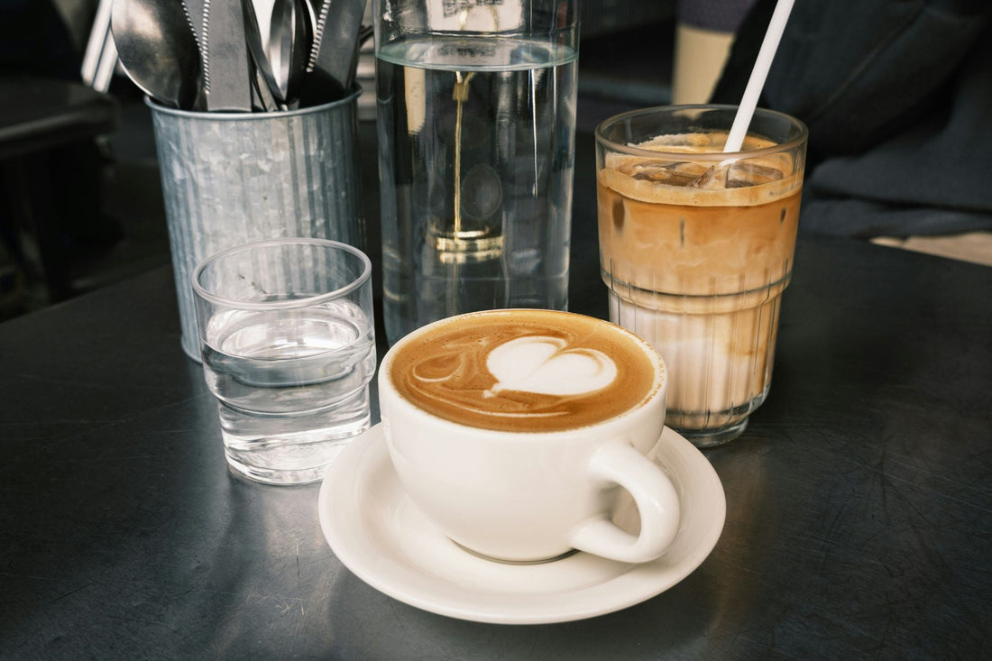 Cup of tea latte with heart-shaped foam art on a table, beside a glass of water and iced tea latte in a café setting