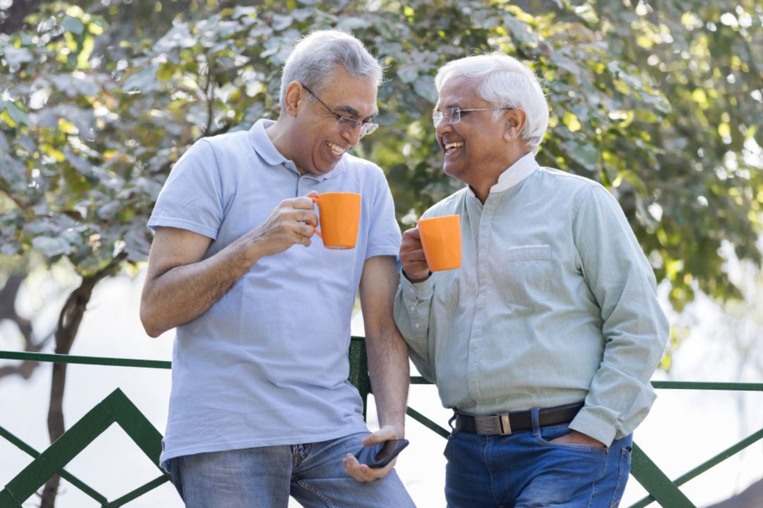 Two mid aged men laughing at each other both holding a cup of tea each in their right hands standing infront of a tree