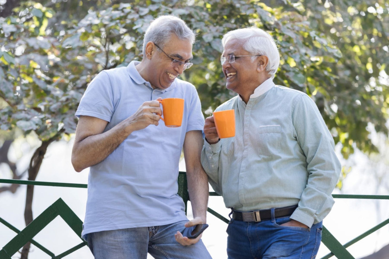 Two mid aged men laughing at each other both holding a cup of tea each in their right hands standing infront of a tree