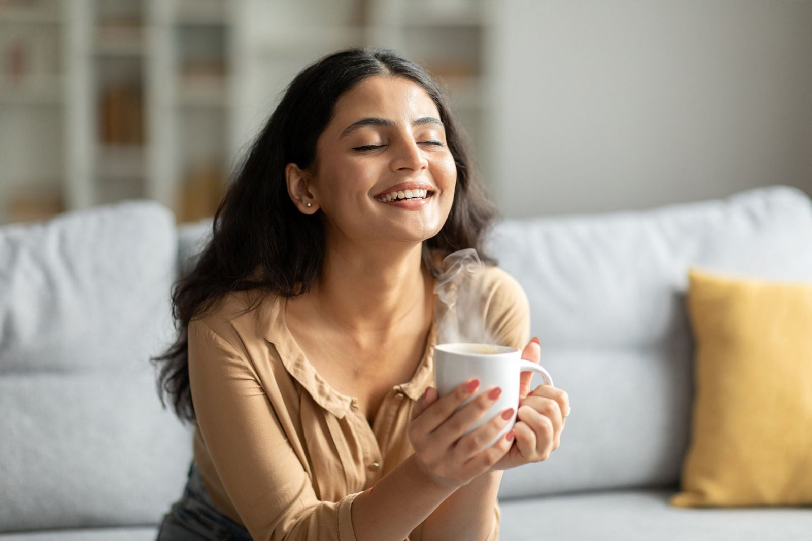 A smiling woman with eyes closed holding a cup of chai with both hands while sitting on a bed