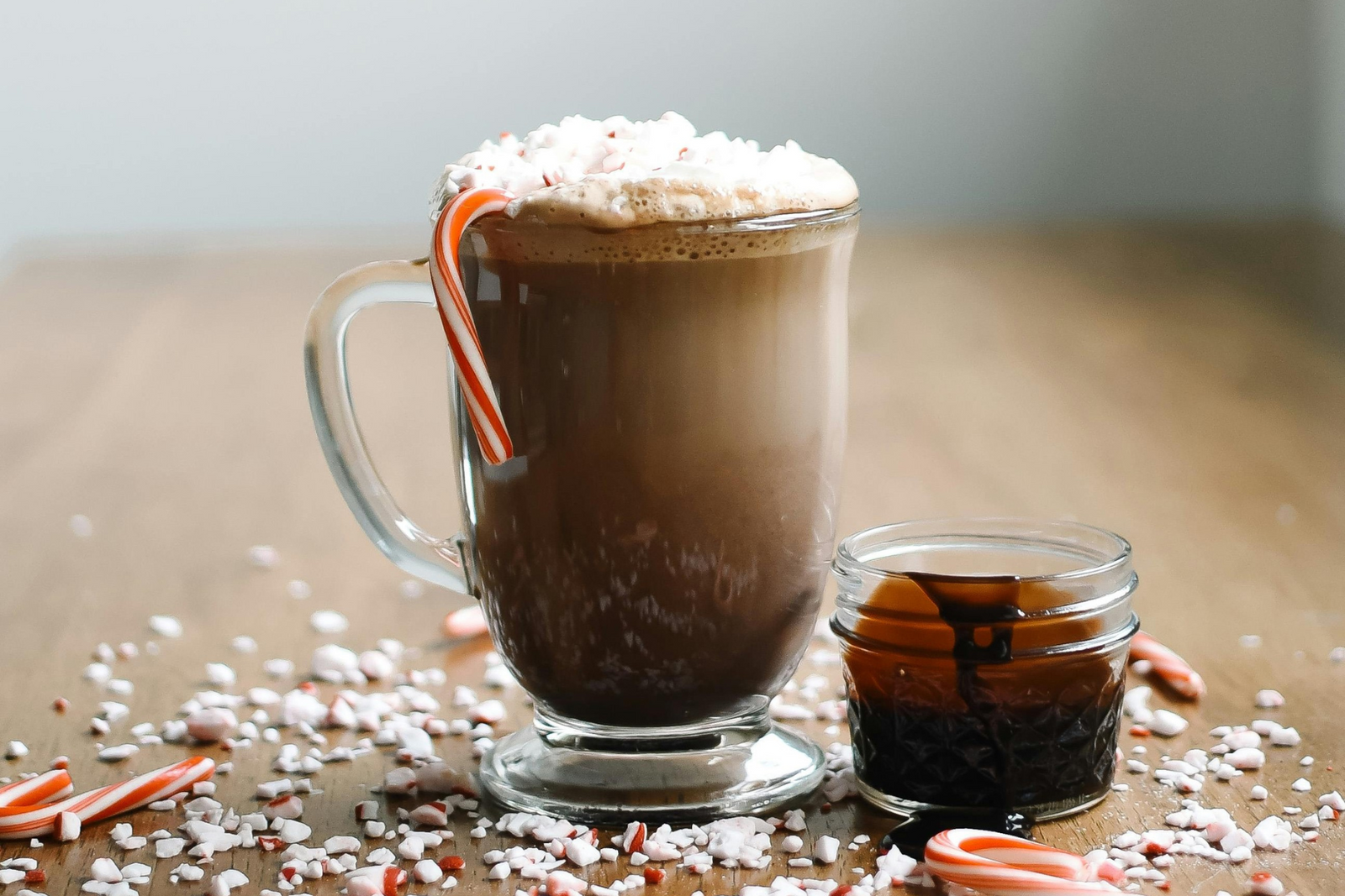Hot chocolate with whipped cream, crushed peppermint, and candy cane in a glass mug, kept next to a small jar of chocolate syrup