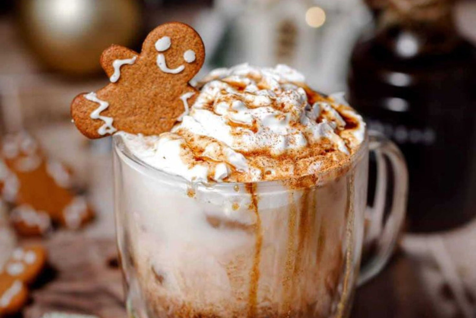 Drink served on a glass mug toppled with whipped cream, drizzled with caramel and a small gingerbread cookie shaped like a smiling gingerbread person perched on the rim of the glass