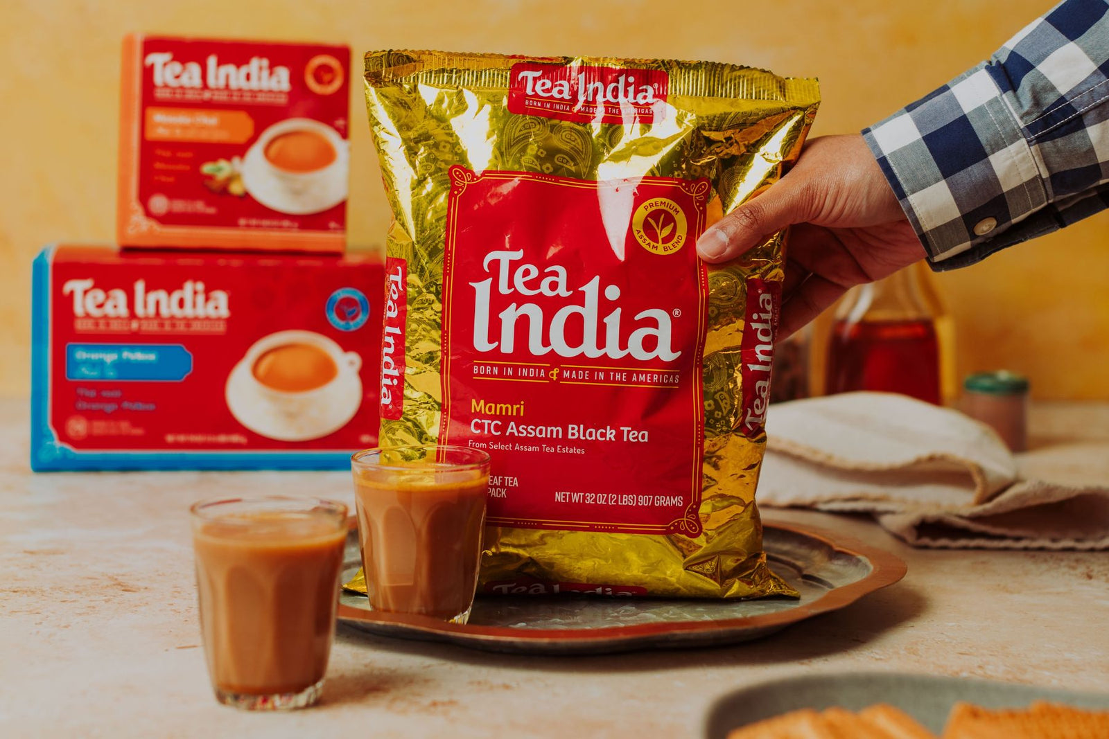 Hand holding a pack of Tea India CTC Assam Black Tea with two glasses of prepared chai with Tea India Orange Pekoe and Masala Chai tea in background