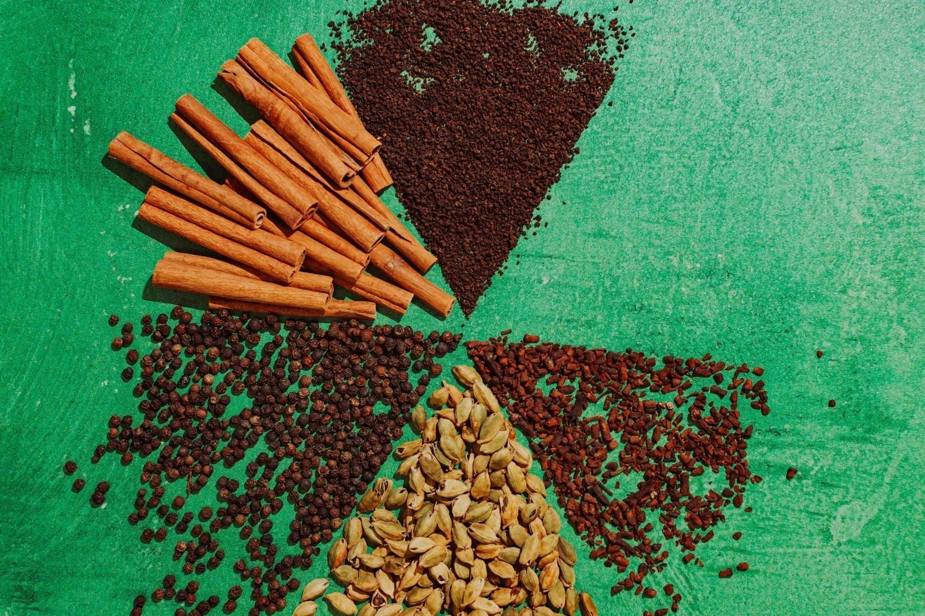 Assorted chai spices and black tea arranged on a green background, including cinnamon sticks, black peppercorns, cardamom pods, cloves, and loose tea leaves.