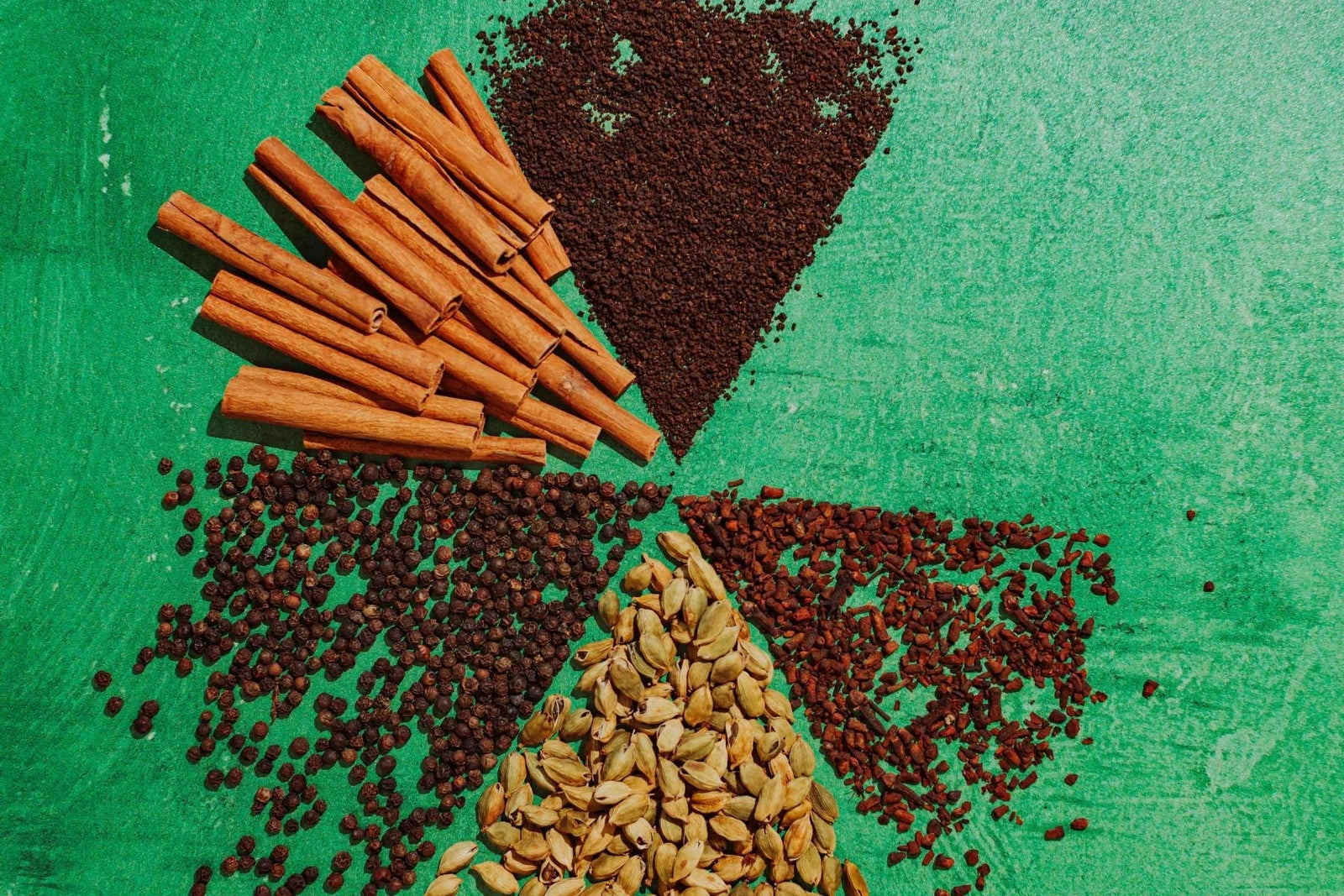 Assorted chai spices and black tea arranged on a green background, including cinnamon sticks, black peppercorns, cardamom pods, cloves, and loose tea leaves.