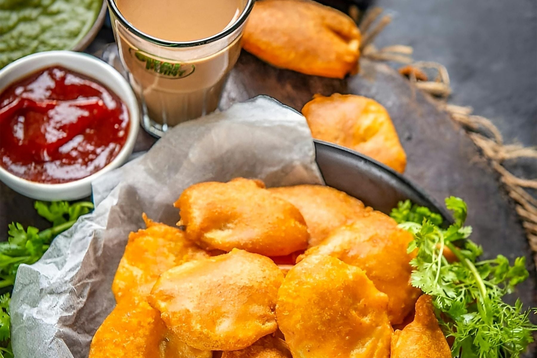 Plate of crispy golden Indian fritters served with green chutney, red sauce, and a glass of chai tea