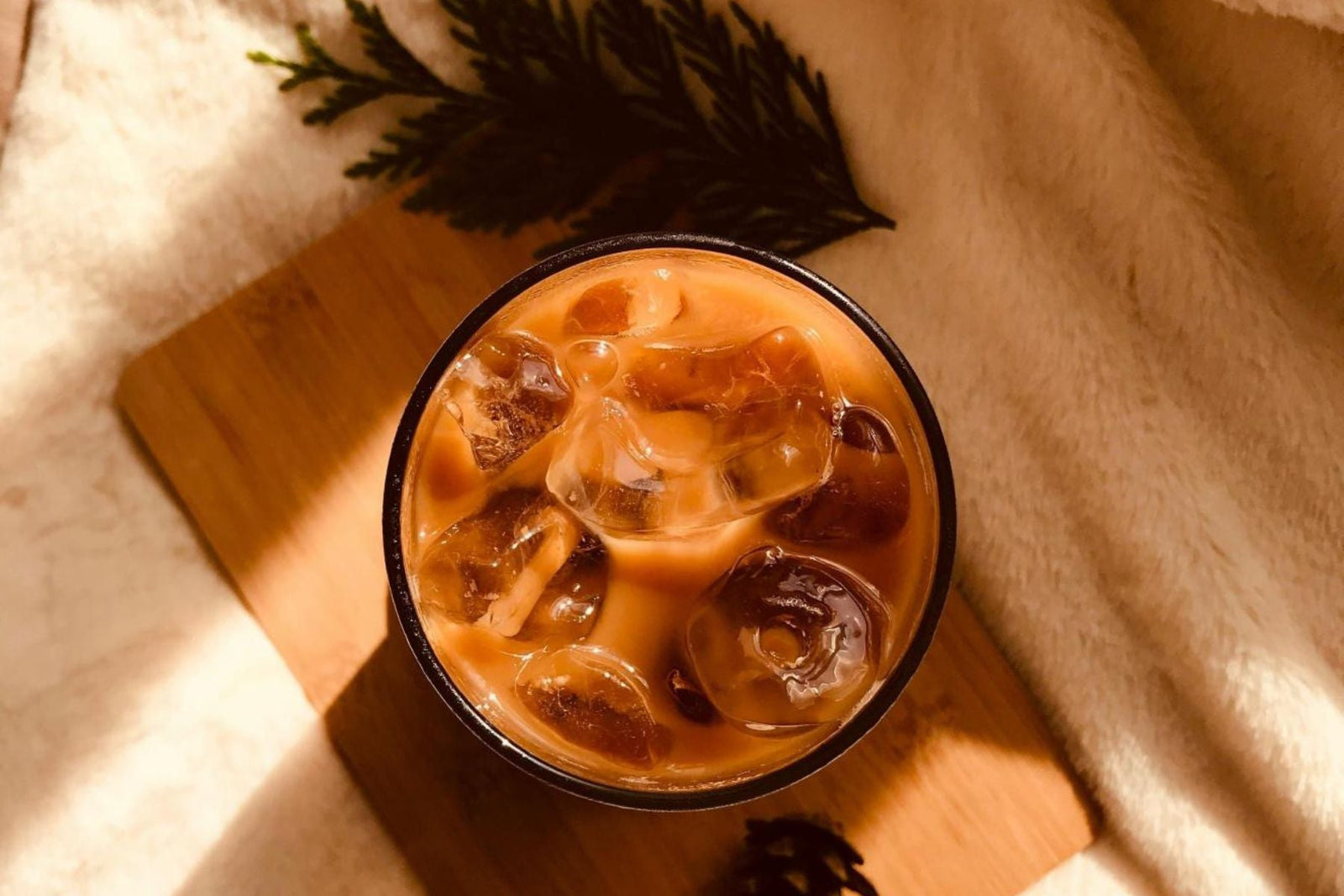 A glass of iced tea with milk and ice cubes on a wooden board, softly lit by warm sunlight.