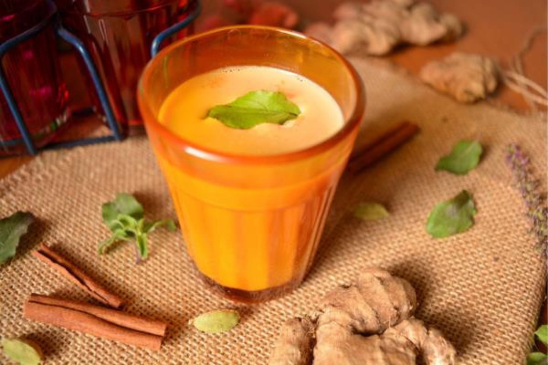 A glass of spiced Indian chai garnished with a mint leaf, surrounded by ginger, cinnamon, and cardamom.