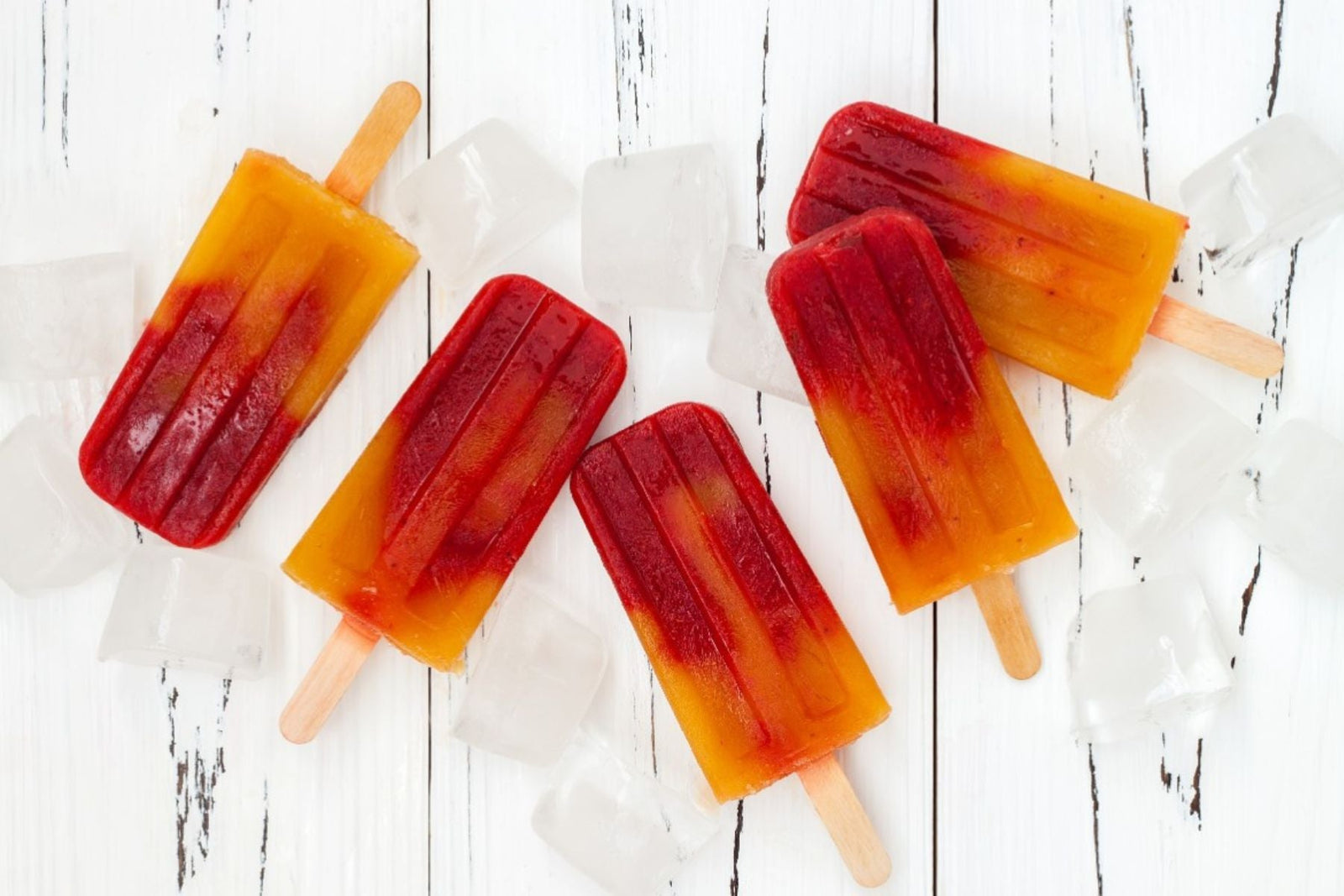 Overhead shot of two-toned red and yellow fruit popsicles scattered with ice cubes.