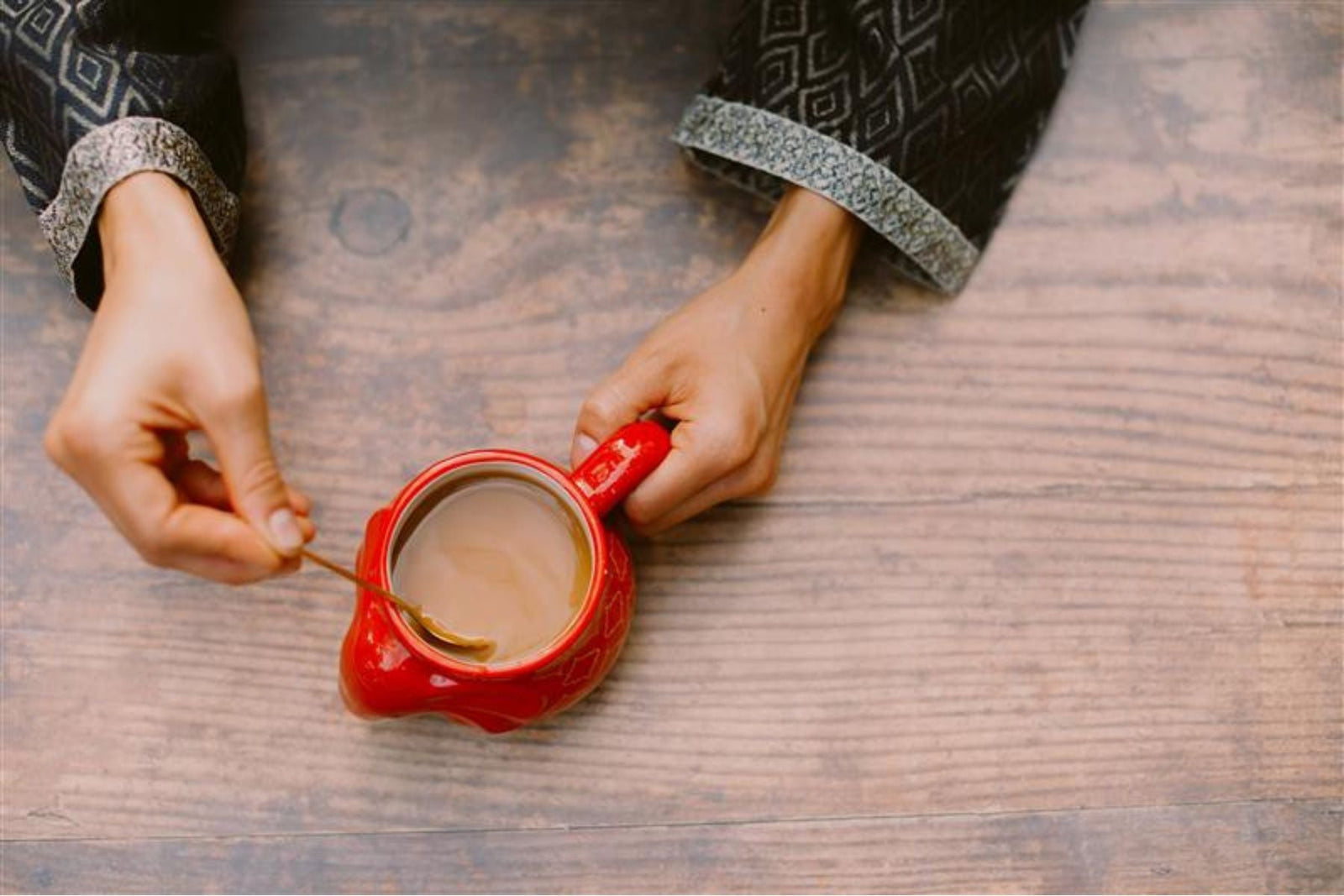 Red color tea mug containing tea and spoon dipped inside it while holding with 2 hands