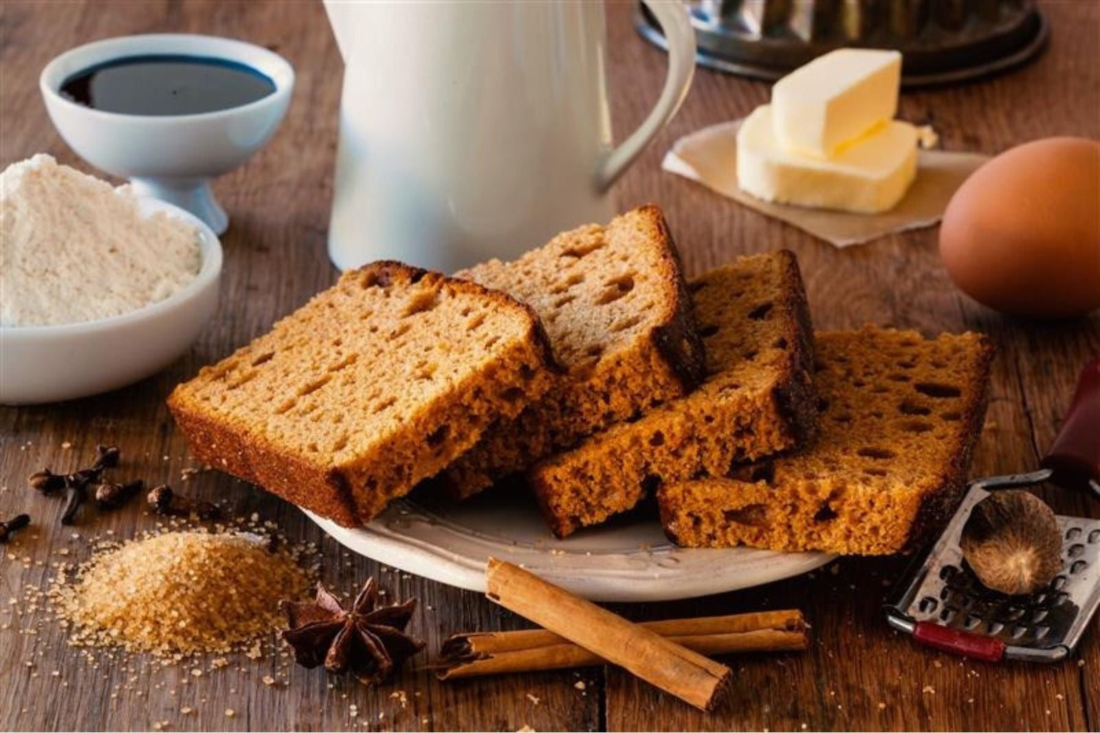 Slices of spiced loaf cake surrounded by raw ingredients like cinnamon and eggs.