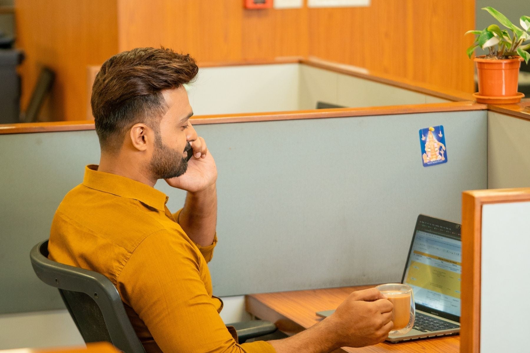Man looking at a laptop sitting in a cubicle holding a cup of tea in his right hand
