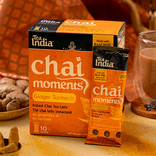 Tea India Chai Moments Ginger Turmeric packaging on a wooden surface with a glass of chai.