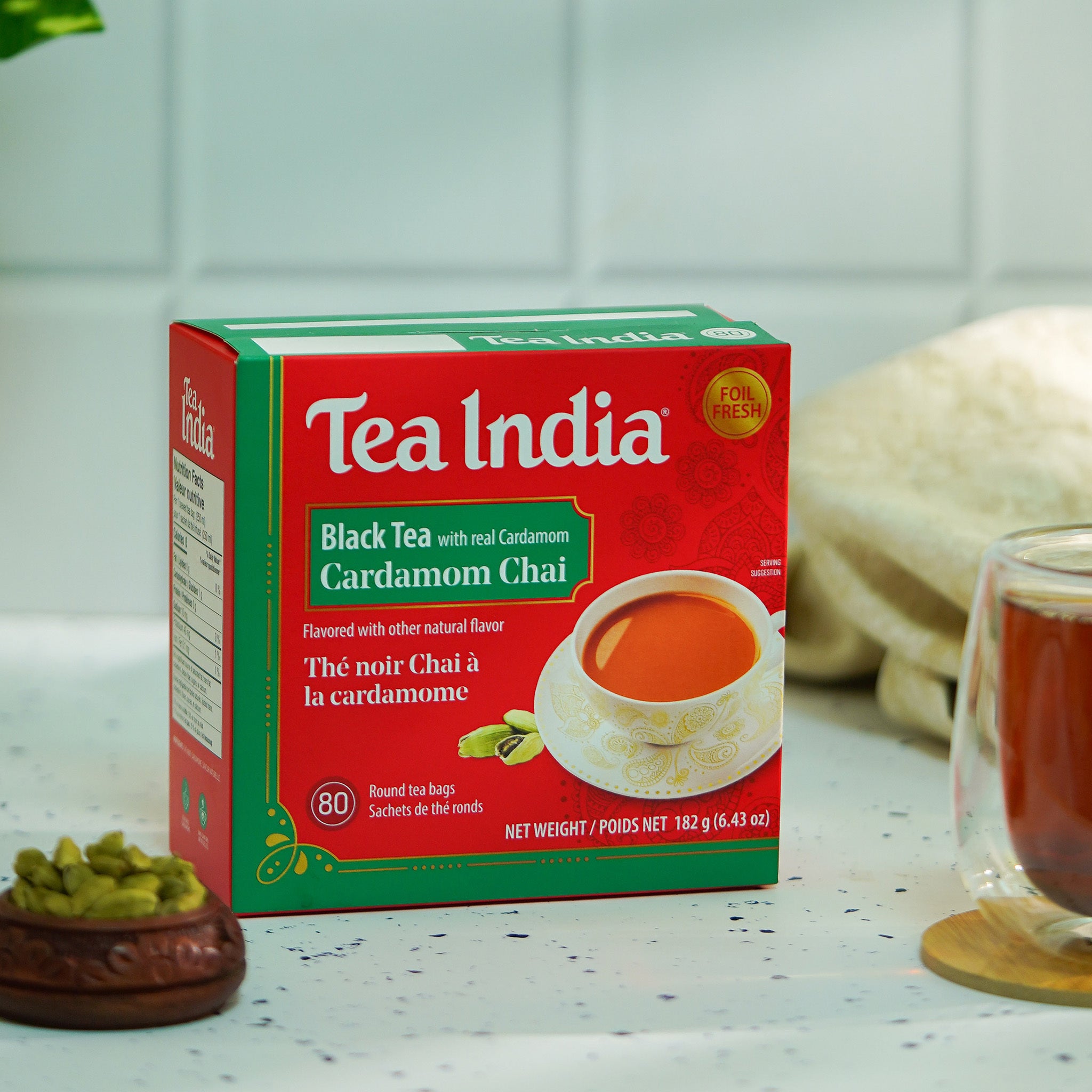 Tea India black tea with real cardamom packaging on a counter next to a cup of tea and a bowl of cardamom pods