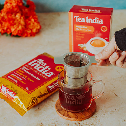 Tea India loose leaf tea packages with a glass mug of tea and a hand holding a tea strainer on a table.