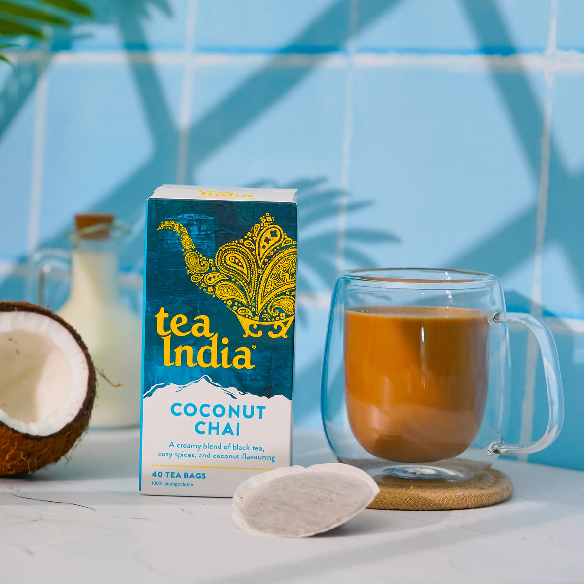 Tea India's Coconut Chai tea bag box on a white counter with a blue tile backsplash. On the counter are a coconut, a round chai tea bag, and a clear glass mug full of prepared chai.