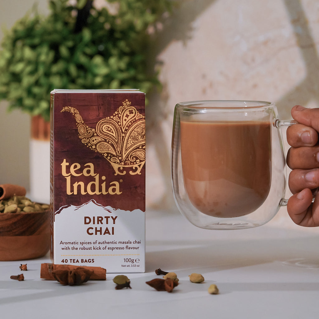 A box of Tea India Dirty Chai tea bags sits on a white counter with spices surrounding it. A hand reaches in from the side of the frame holding a clear glass mug full of prepared chai.