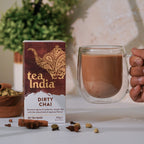 A box of Tea India Dirty Chai tea bags sits on a white counter with spices surrounding it. A hand reaches in from the side of the frame holding a clear glass mug full of prepared chai.