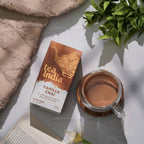 A box of tea India Vanilla Chai tea bags sits next to a glass mug filled with brewed chai on a light surface, styled with a beige towel, green leaves, and an open book.