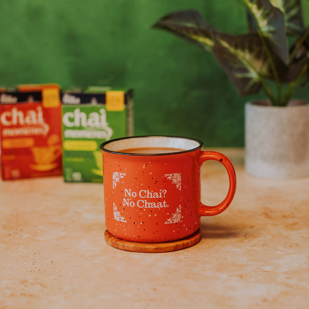 an orange campfire mug that says "No Chai? No Chaat." in front of boxes of Tea India, a houseplant, and a green background