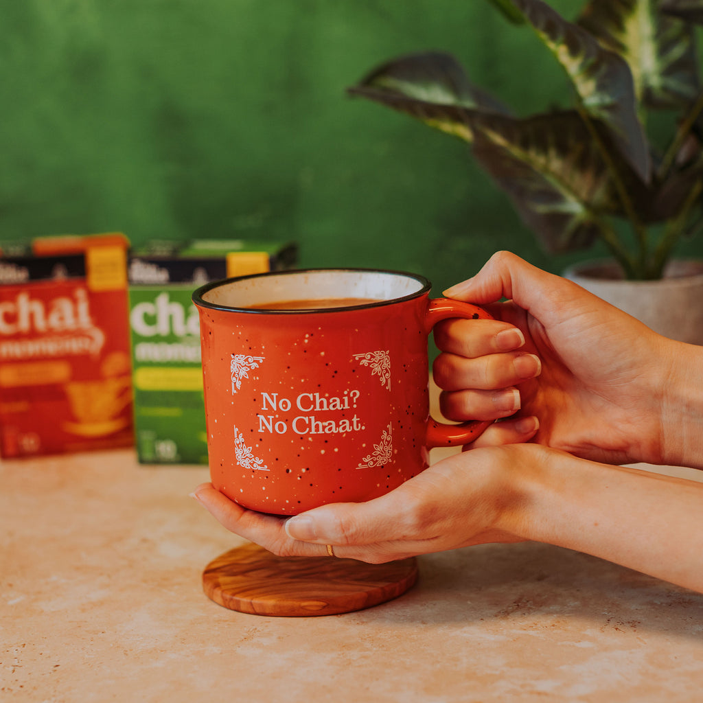 hands holding an orange campfire mug that says "No Chai? No Chaat." in front of boxes of Tea India, a houseplant, and a green background