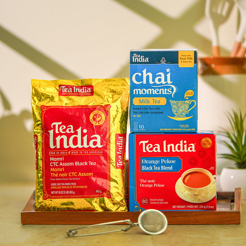 Three Tea India tea products arranged on a wooden tray: a large gold bag of Mamri CTC Assam Black Tea, a blue box of Chai Moments Milk Tea, and a red box of Orange Pekoe Black Tea Blend. A small metal tea strainer sits in front on a light surface.