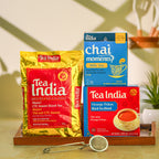 Three Tea India tea products arranged on a wooden tray: a large gold bag of Mamri CTC Assam Black Tea, a blue box of Chai Moments Milk Tea, and a red box of Orange Pekoe Black Tea Blend. A small metal tea strainer sits in front on a light surface.