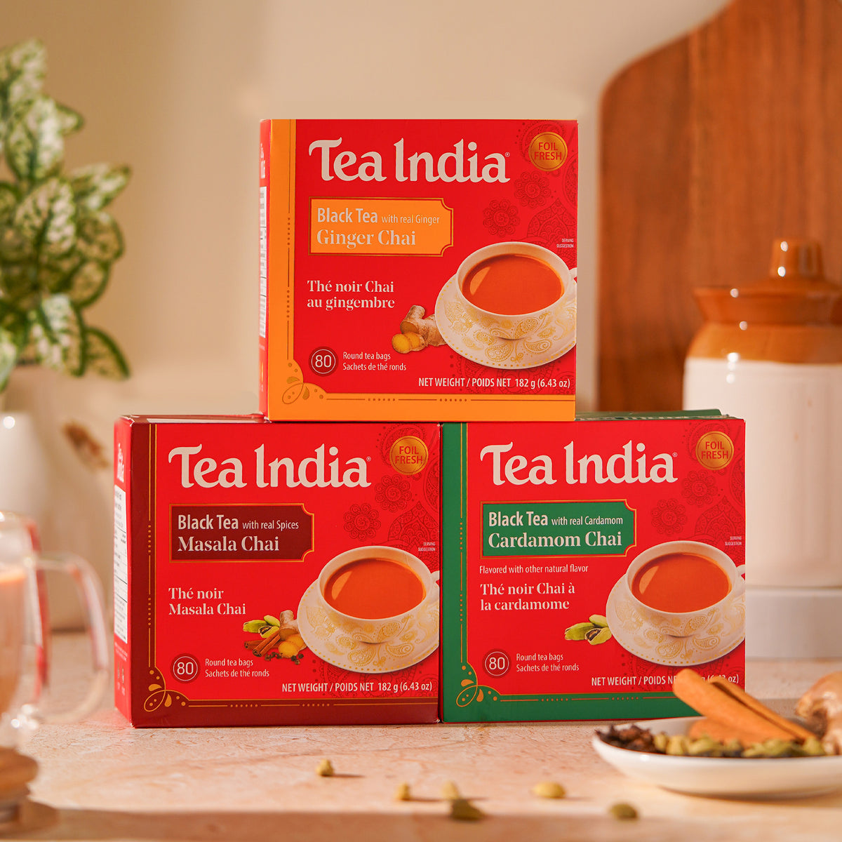 Three boxes of Tea India black tea chais arranged on a tabletop: a Ginger Chai box on top, a Masala Chai box on the left, and a Cardamom Chai box on the right. The boxes are red with different flavor panels, set in a warm kitchen-like environment with a glass of tea, spices, and a ceramic jar in the background.