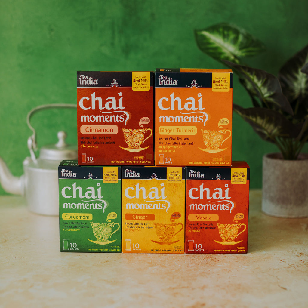 Five boxes of The India Chai Moments tea mix arranged on a light stone surface against a green wall, with a white teapot on the left and a potted plant on the right.