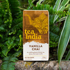 A box of Tea India Vanilla Chai tea bags standing upright outdoors on a stone surface, surrounded by green fern leaves and plants.