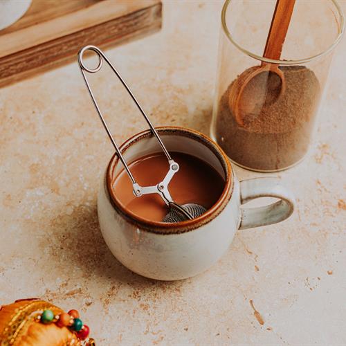 A stainless steel tea infuser steeping loose leaf tea inside a white ceramic mug filled with hot chai
