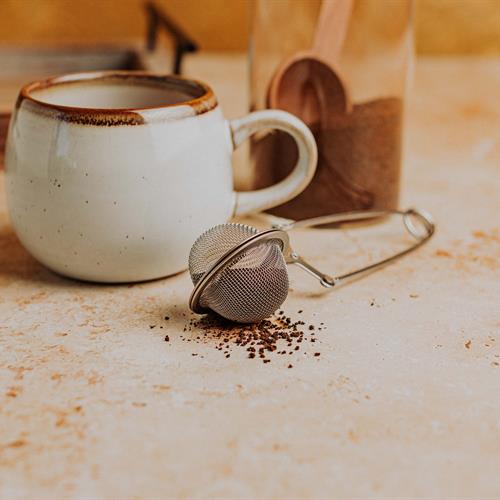 A tea infuser next to a pile of loose tea leaves, with a ceramic mug and jar of tea in the background