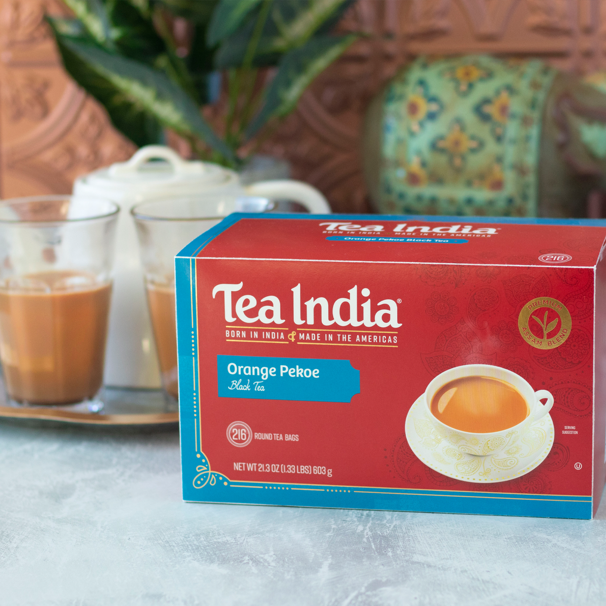 A box of Tea India Orange Pekoe Black Tea on a counter, with two glasses of tea and a white teapot in the blurred background.