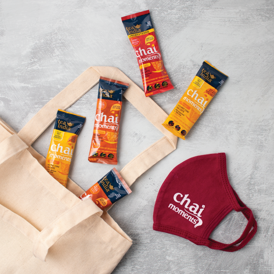 A beige tote bag with several &quot;chai moments&quot; instant chai packets and a branded maroon cloth face mask