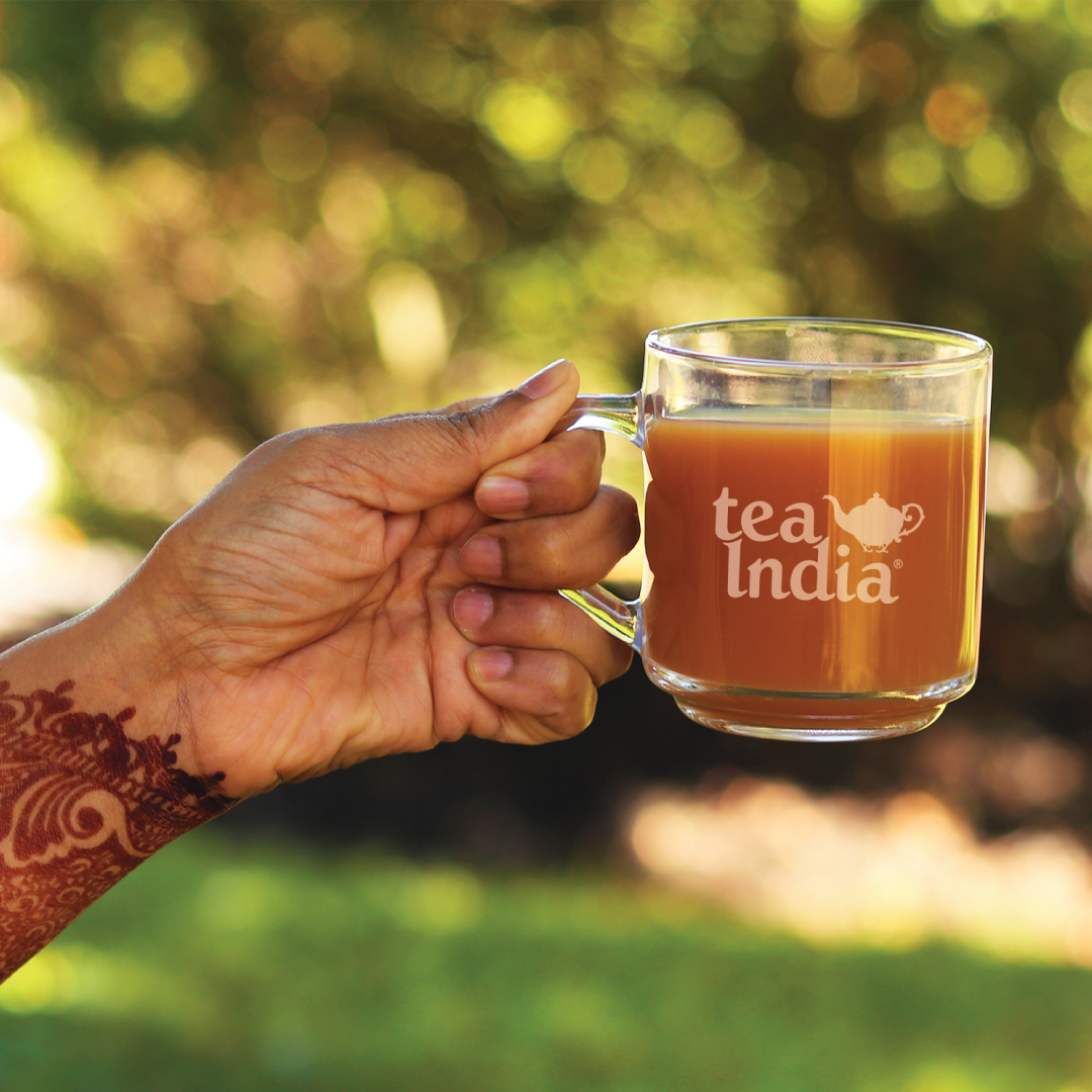 A hand with a henna design holding a clear glass Tea India branded mug filled with hot chai tea, with a blurred outdoor background